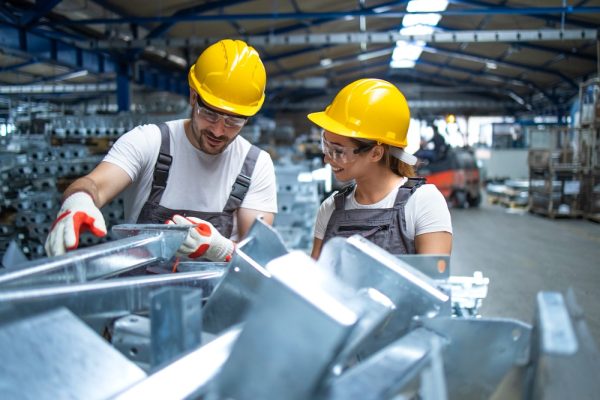 Trabajadores con casco de seguridad revisando piezas metálicas en planta de fabricación industrial.