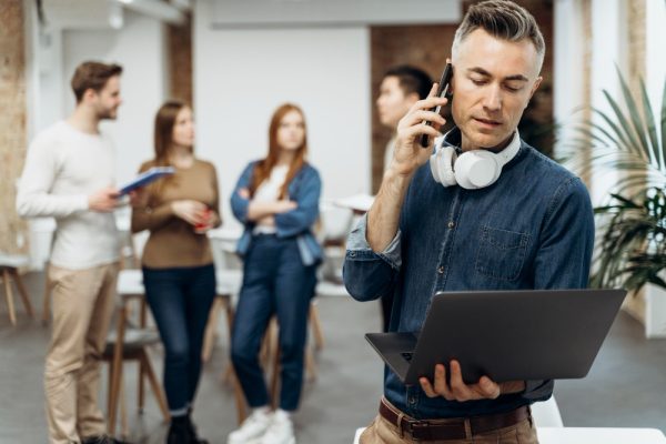 Ejecutivo hablando por teléfono mientras trabaja con un portátil, con equipo colaborando en segundo plano.