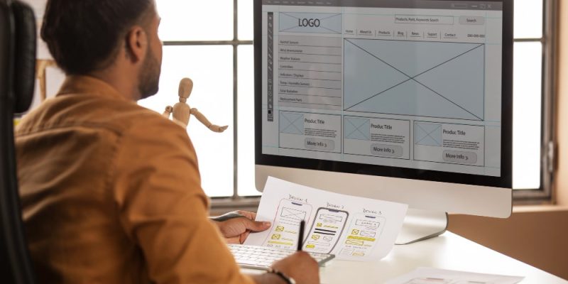 "Profesional diseñando un sitio web con prototipos en papel y maqueta digital en monitor.