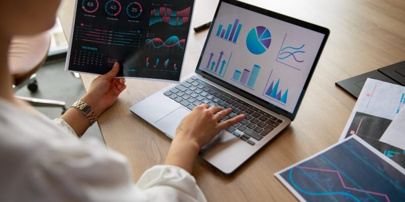 Persona analizando gráficos financieros en laptop y hojas impresas sobre escritorio.