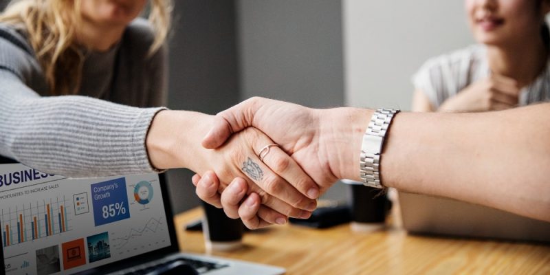 Dos personas estrechando la mano en acuerdo empresarial con gráficos de crecimiento en pantalla.